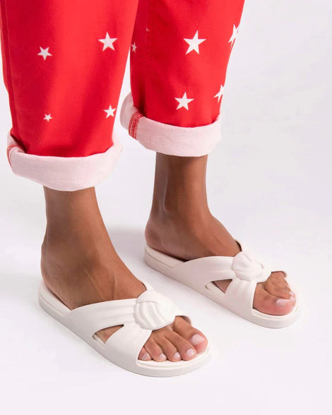 A model's legs in red patterned pants and a pair of white Melissa Plush slides with a twist front strap detail.
