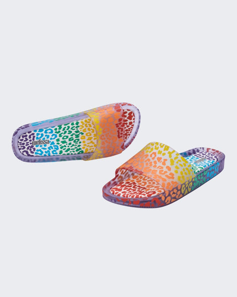 An angled front and top view of a pair of clear / orange Melissa Rainbow Beach slides with a clear base and a rainbow cheetah print pattern.