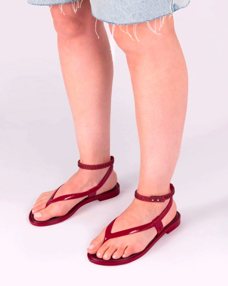 A model's legs in denim shorts, wearing a pair of red Melissa Unique sandals with flip flop straps and a woven detail ankle strap.