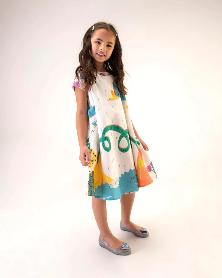 A young model posing for a picture in a patterned dress and a pair of silver Mini Melissa Sweet Love Princess flats, with a silver base, Cinderella in script on the side, a heart detail on the toe, and a drawing of Cinderella on the sole.
