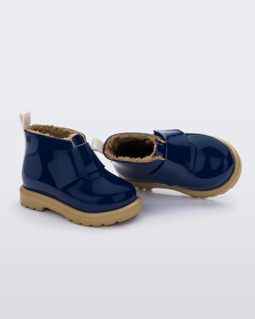 An angled top and side view of a pair of blue/beige Mini Melissa Chelsea blue boots with a velcro strap, dark blue base, beige sole and beige fuzzy lining.