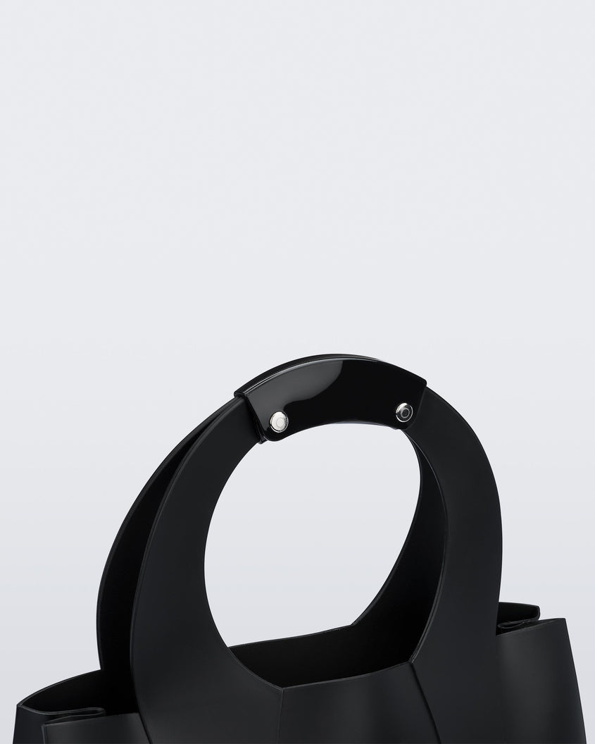 A zoomed in view of the top of a black Melissa Essential Tropicana bag with a circular handle with two silver studs.