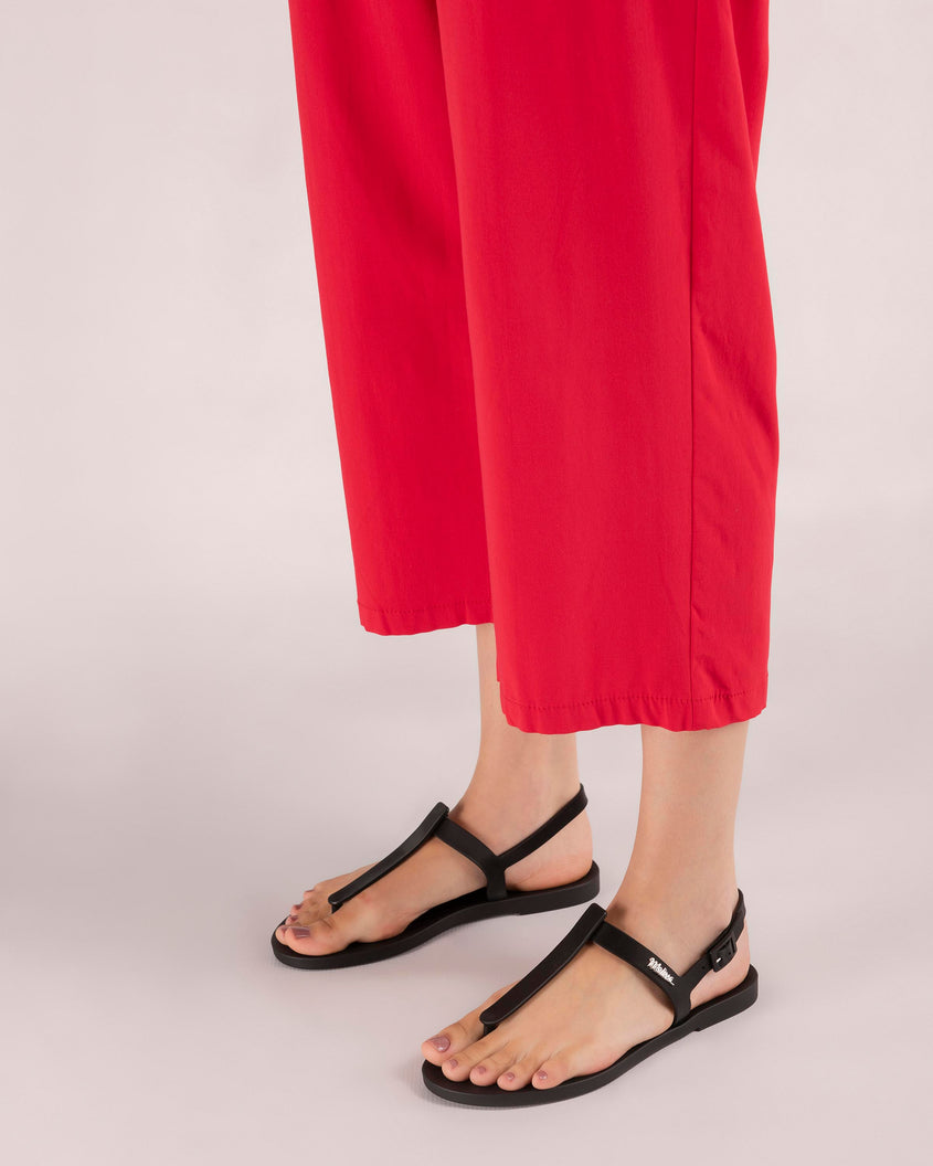 A model's legs in red pants, wearing a pair of black Melissa Sun Ventura sandals with a top strap, reading melissa in silver, intersected by a vertical front strap detail and an ankle strap.