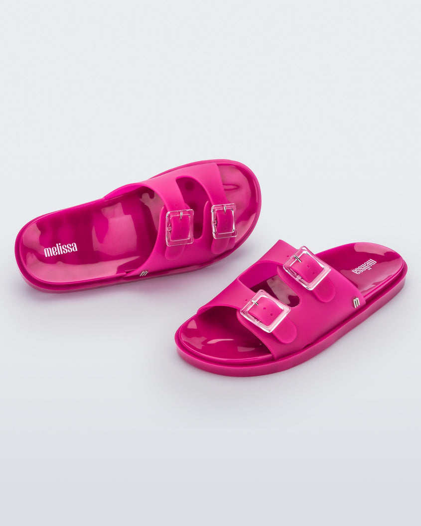 An angled front and top view of a pair of dark pink Melissa Wide Slides with two straps fastened at the top with two clear buckles.