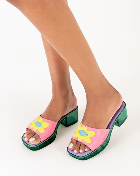 A model's legs wearing a pair of Melissa Shape slides with green platform heel, a pink wide front strap with a yellow and blue flower on the top of the strap and a purple insole
