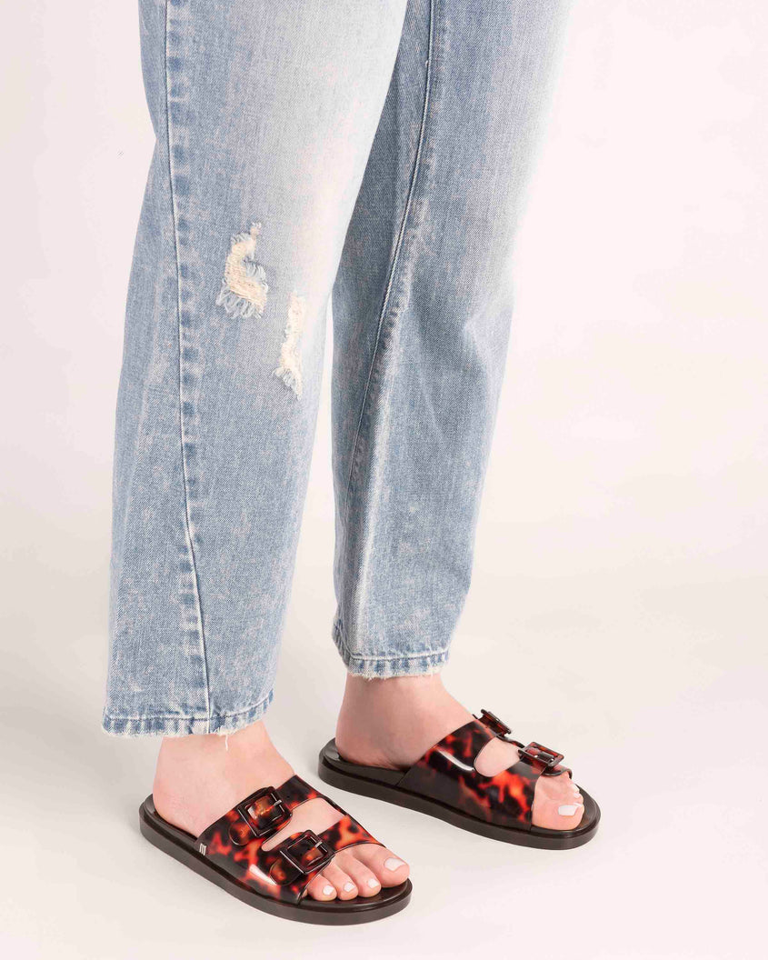 A model's legs wearing a pair of Melissa Wide slide sandals with brown sole and Brown Tortoise colored straps