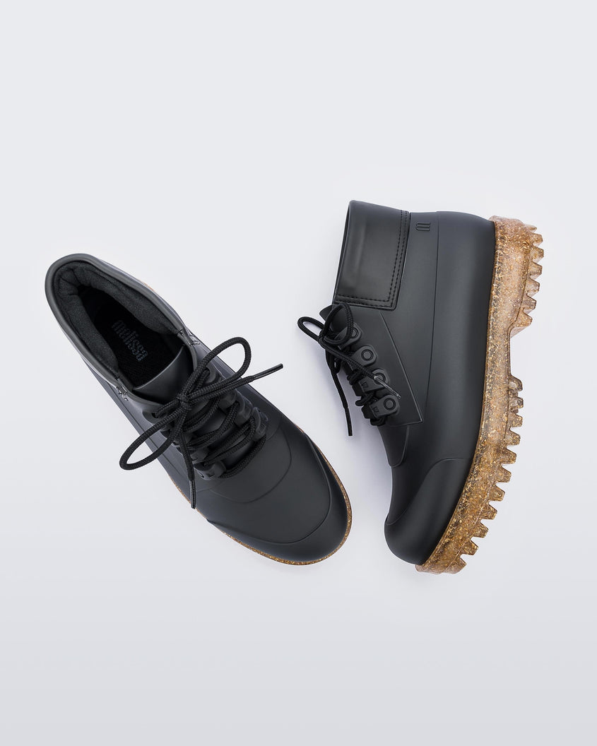 A top and side view of a pair of matte black / rice husk Melissa Ares combat boots with a black sole, black laces and brown / gold glitter rice husk tractor sole.