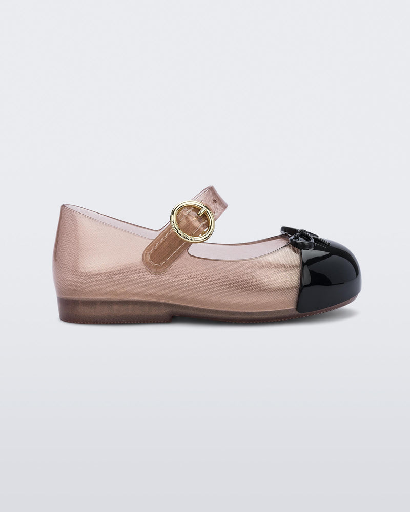 SIde view of the Mini Melissa Sweet Love ballet flat with buckle strap. The body of the shoe is a transparent rose gold color, the shoe has a black cap to with a 3D bow applique on the top of the toe. The buckle hardware on the strap is gold.