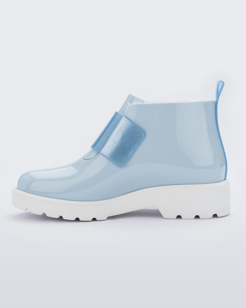 An inner side view of a blue Mini Melissa Chelsea boot with a light blue base, glitter blue velcro front strap and a white sole.
