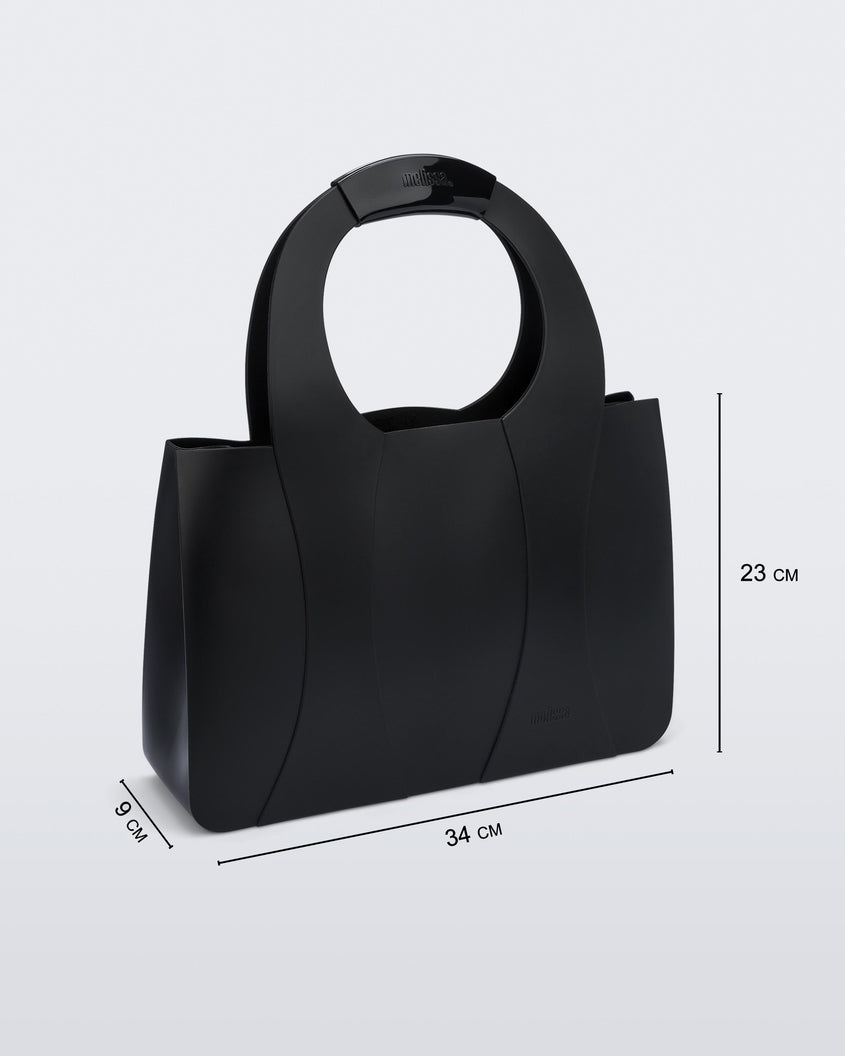 An angled front view of a black Melissa Essential Tropicana bag with a circular handle that has the word melissa on it, showing the dimensions of the bag which read 9 centermeters deep, 34 centermeters wide and 23 centermeters high.