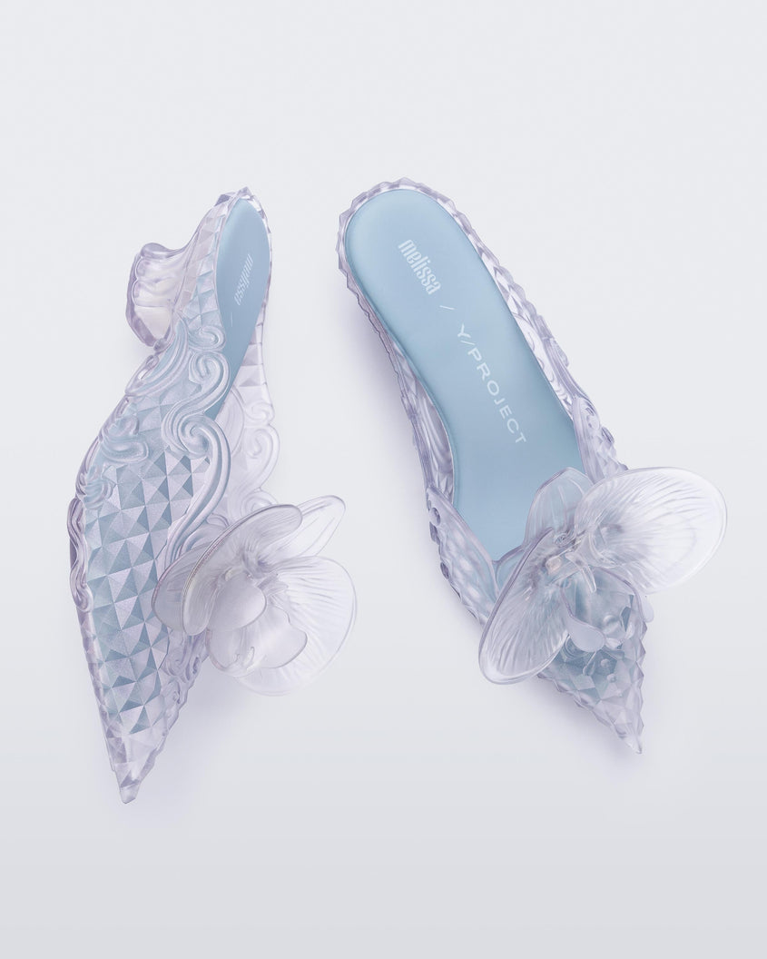 Top and side view of pair of Melissa Flower Court mules in clear with kitten heel and a 3D flower above the toe. The top view shows the light blue colored insole