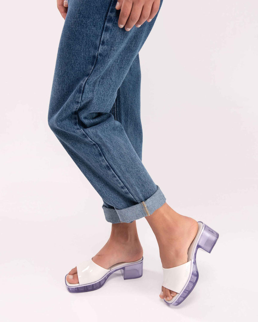 A model's legs wearing a pair of Melissa Shape slides with clear heel and white wide strap