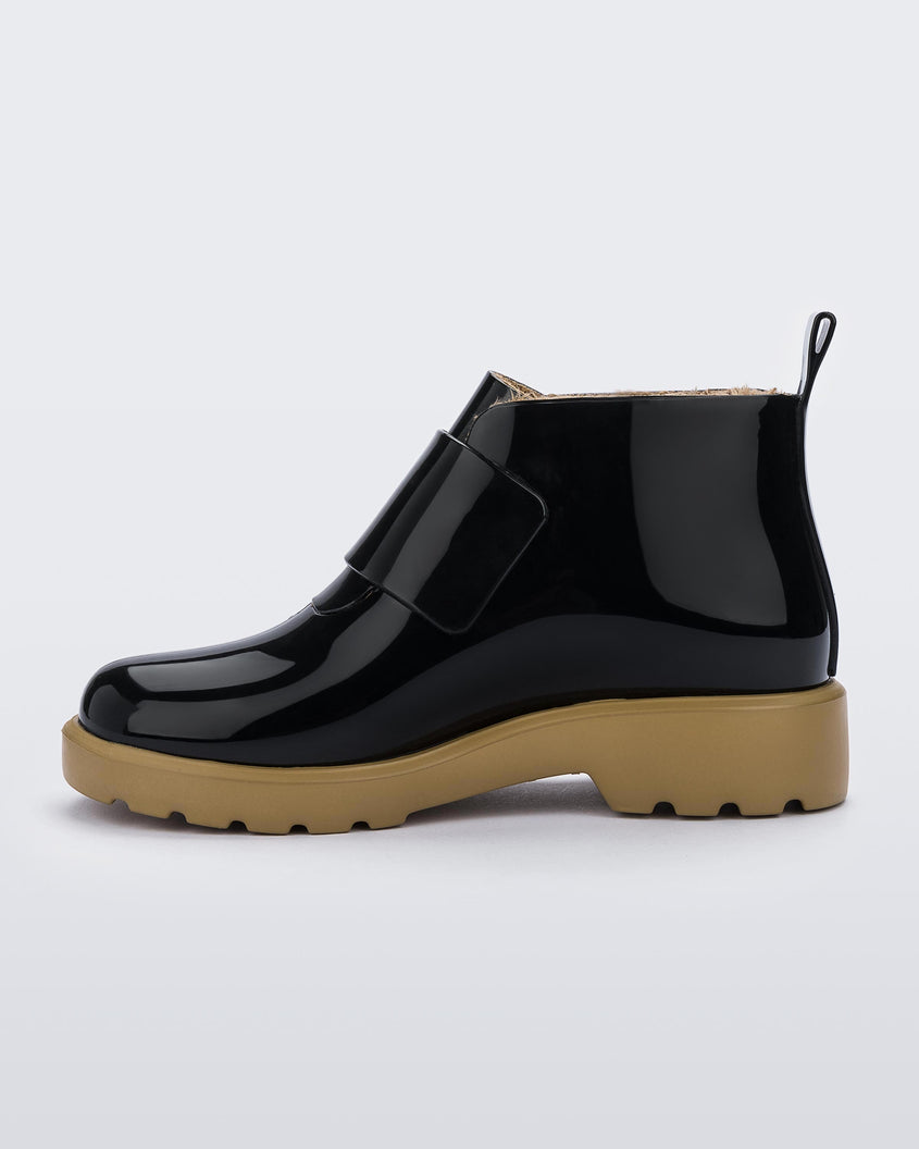 An inner side view of a black/beige Mini Melissa Chelsea boot with a black base, black velcro front strap and beige sole.