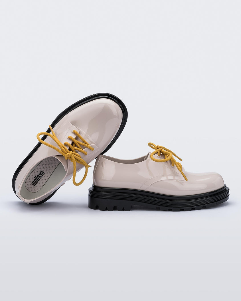 Two sneakers leaning on eachother: A side and top view of a pair of beige Melissa Bass sneakers with a beige base, yellow laces and a black sole.