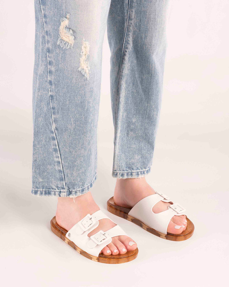 Model's legs wearing a pair of Melissa Wide slide sandals with gold glitter sole and white double straps with buckles