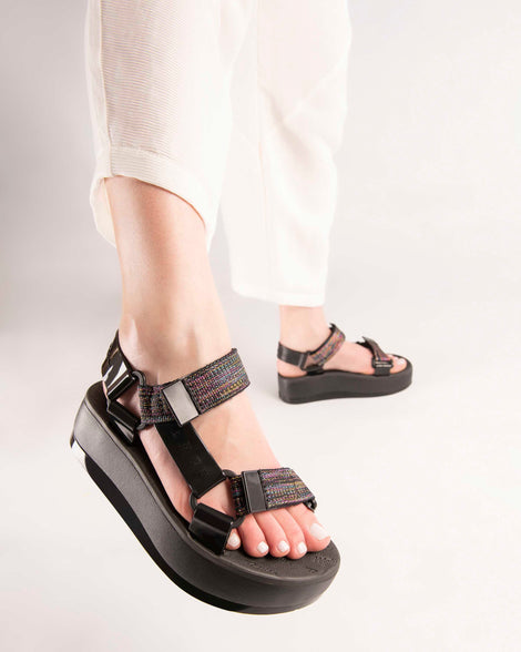 A model's legs wearing tan pants and a pair of black Melissa Papete Platform sandals with a black multicolor straps.