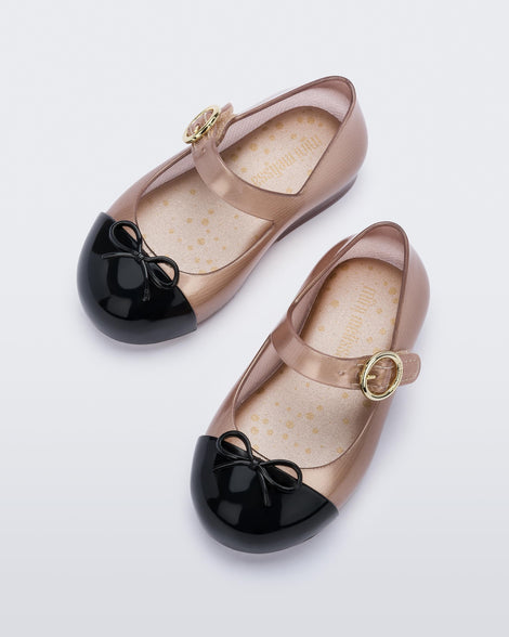 Top view of the Mini Melissa Sweet Love ballet flats with buckle strap. The body of the shoe is a transparent rose gold color, the shoe has a black cap to with a 3D bow applique on the top of the toe. The buckle hardware on the strap is gold.