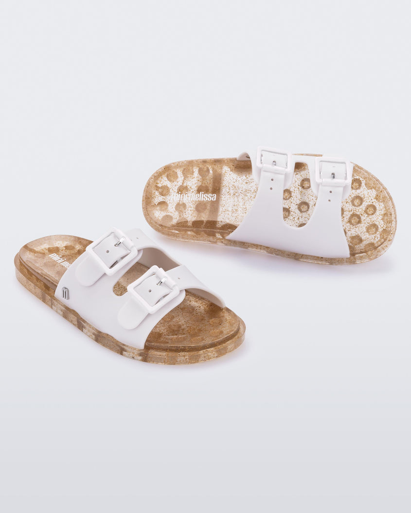 An angled front and top view of a pair of Beige Mini Melissa Wide Slides with a white top with two white buckles and a beige sole.