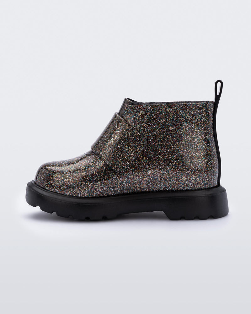 An inner side view of a black/glitter multicolor Mini Melissa Chelsea boot with a multicolor glitter base, velcro front strap and a black sole.