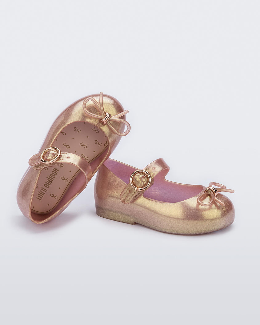 An angled side and top view of a pair of pink Mini Melissa Sweet Love flats with a top strap and a bow detail on the toe, leaning on one another.