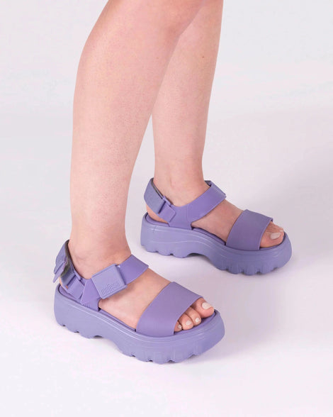 Close up shot of a model's legs, they are wearing a pair of the Melissa Kick Off platform sandals in lilac. The sandal has an ankle strap with a plastic clasp closure on the front ankle side, additional velcro adjustable strap is on the back of the ankle strap.