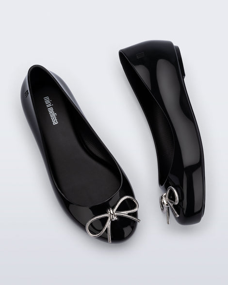Side and top view of a pair of black Sweet Love Chrome kids flats with metallic silver bow