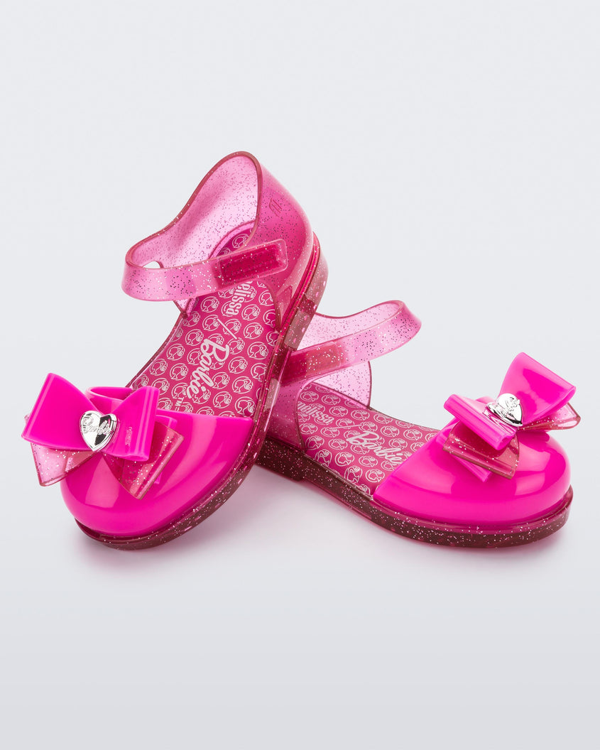 An angled top and side view of a pair of glitter pink Mini Melissa sandals with a Barbie bow detail on the front toe, pink glitter ankle strap and a Barbie logo sole