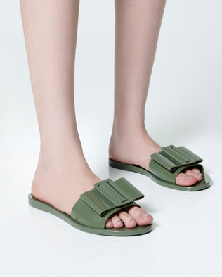 Model's legs  wearing a pair of dark green Babe slide sandals with large bow on top.