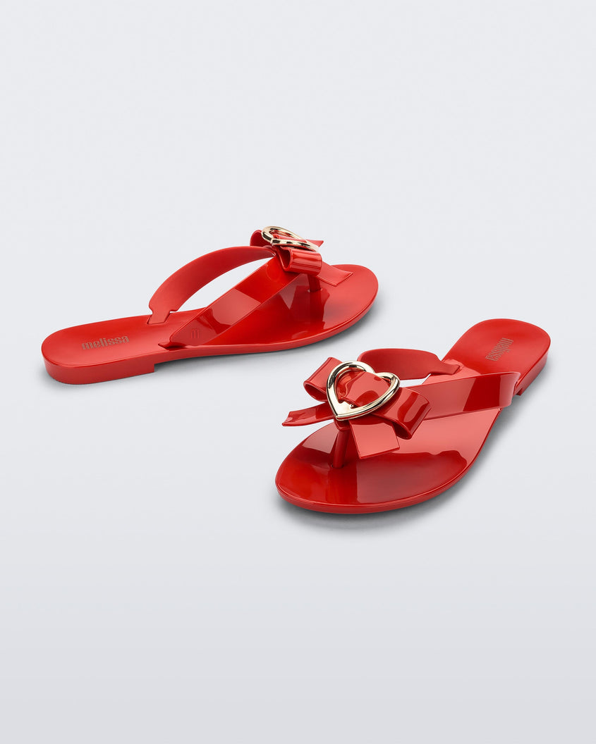 An angled front and side view of a pair of red Melissa Harmonic Heart flip flops with a red bow and gold heart detail on the straps