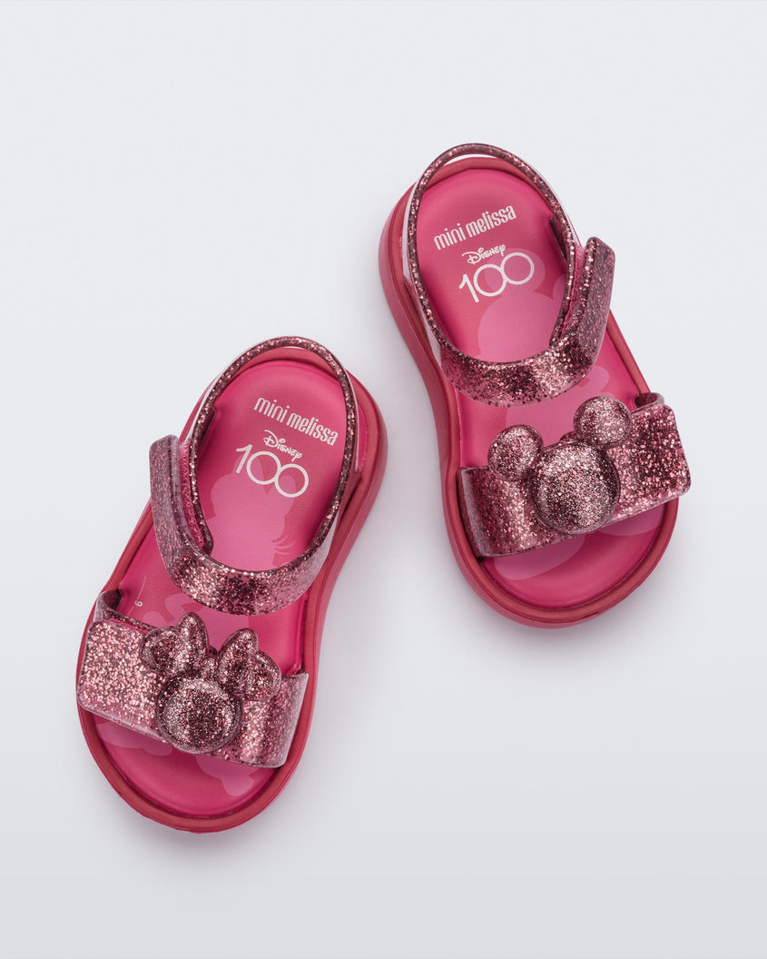 A top view of a pair of glitter pink Mini Melissa Jump sandals with a Mickey Mouse logo detail on the front strap and an ankle strap