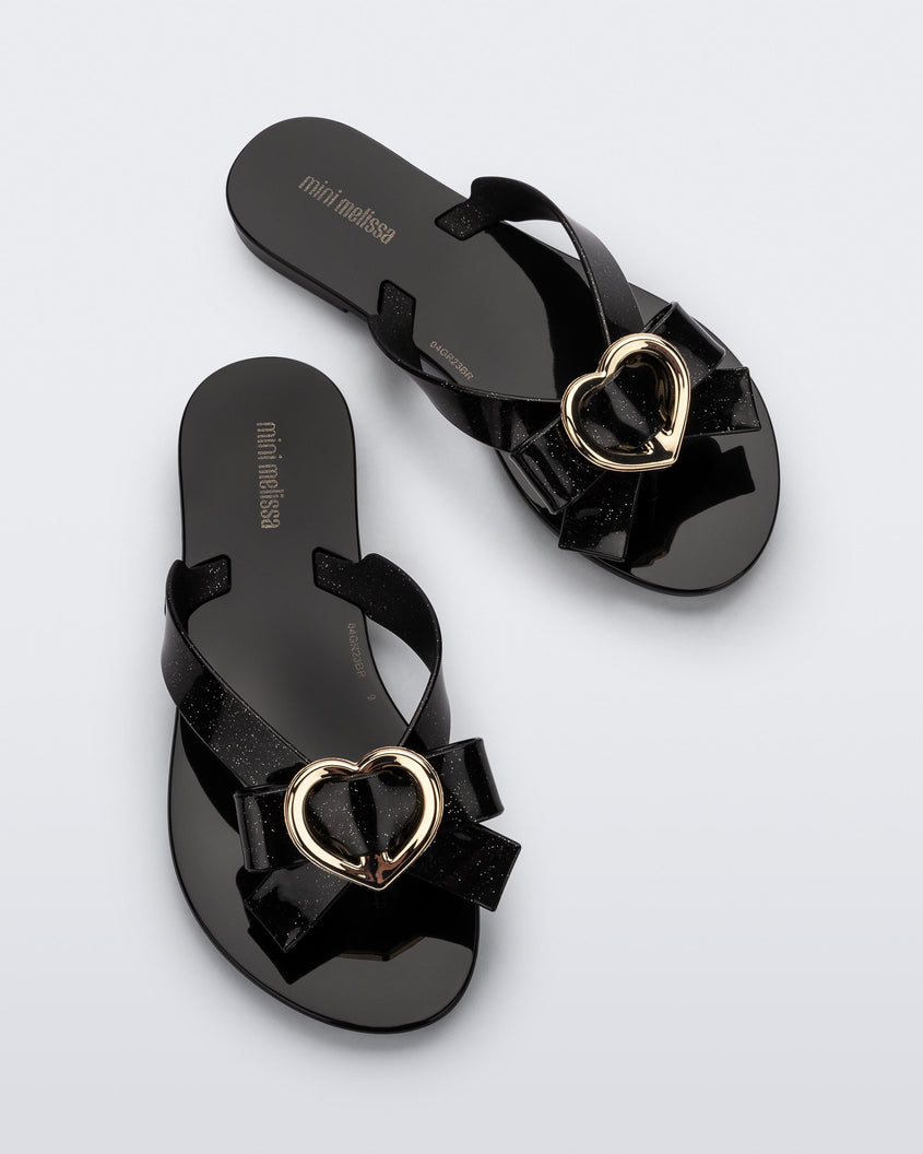 Top view of a pair of black Mini Melissa Harmonic Heart flip flops with a black bow and gold heart detail on the straps