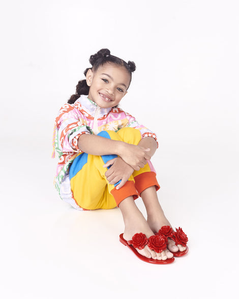 Child model in a multicolor shirt and yellow pants wearing of a pair of a black Harmonic Springtime kids flip flop with three beige flowers.