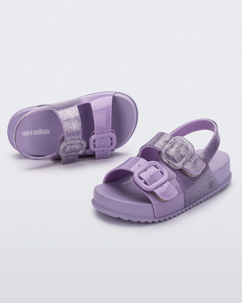 An angled top and side view of a pair of lilac glitter Mini Melissa Cozy sandals with with two front straps with buckle detail