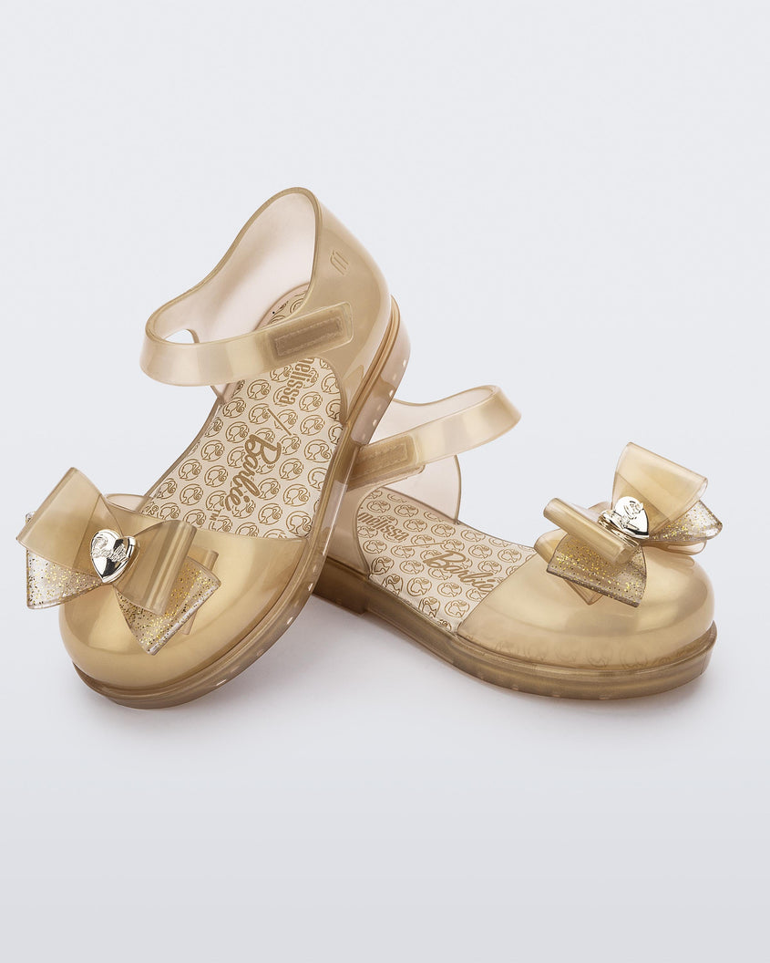 A top view of a pair of pearly beige Mini Melissa sandals with a Barbie bow detail on the front toe, pearly beige ankle strap and a Barbie logo sole