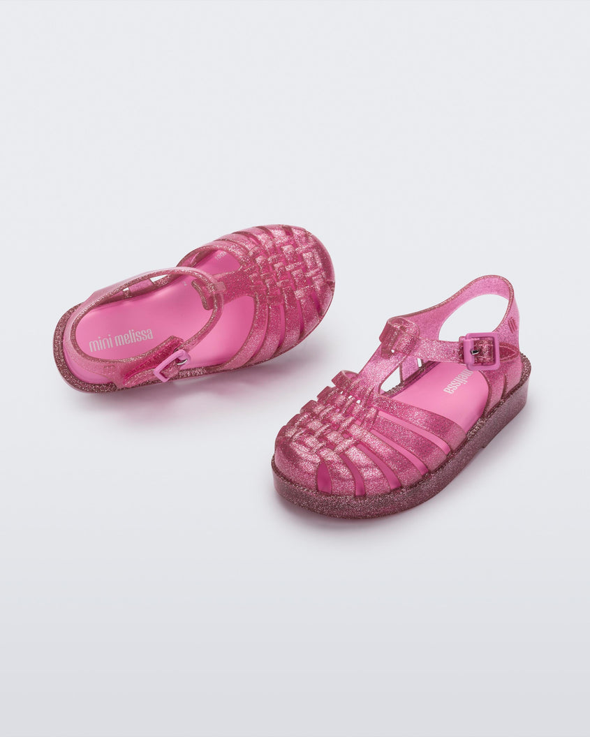 Angled view of a pair of Mini Melissa Possession baby sandals in glitter pink with velcro buckle closure on the ankle straps