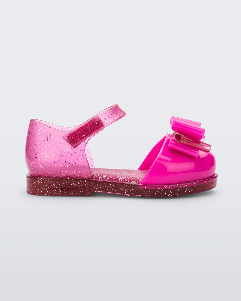 Side view of a glitter pink Mini Melissa sandal with a Barbie bow detail on the front toe, pink glitter ankle strap and a Barbie logo sole