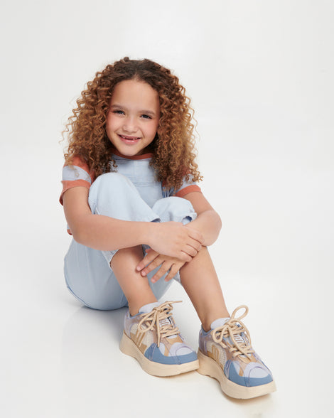Child model sitting down in blue overalls wearing a pair of blue X Beat kids sneakers with beige trim