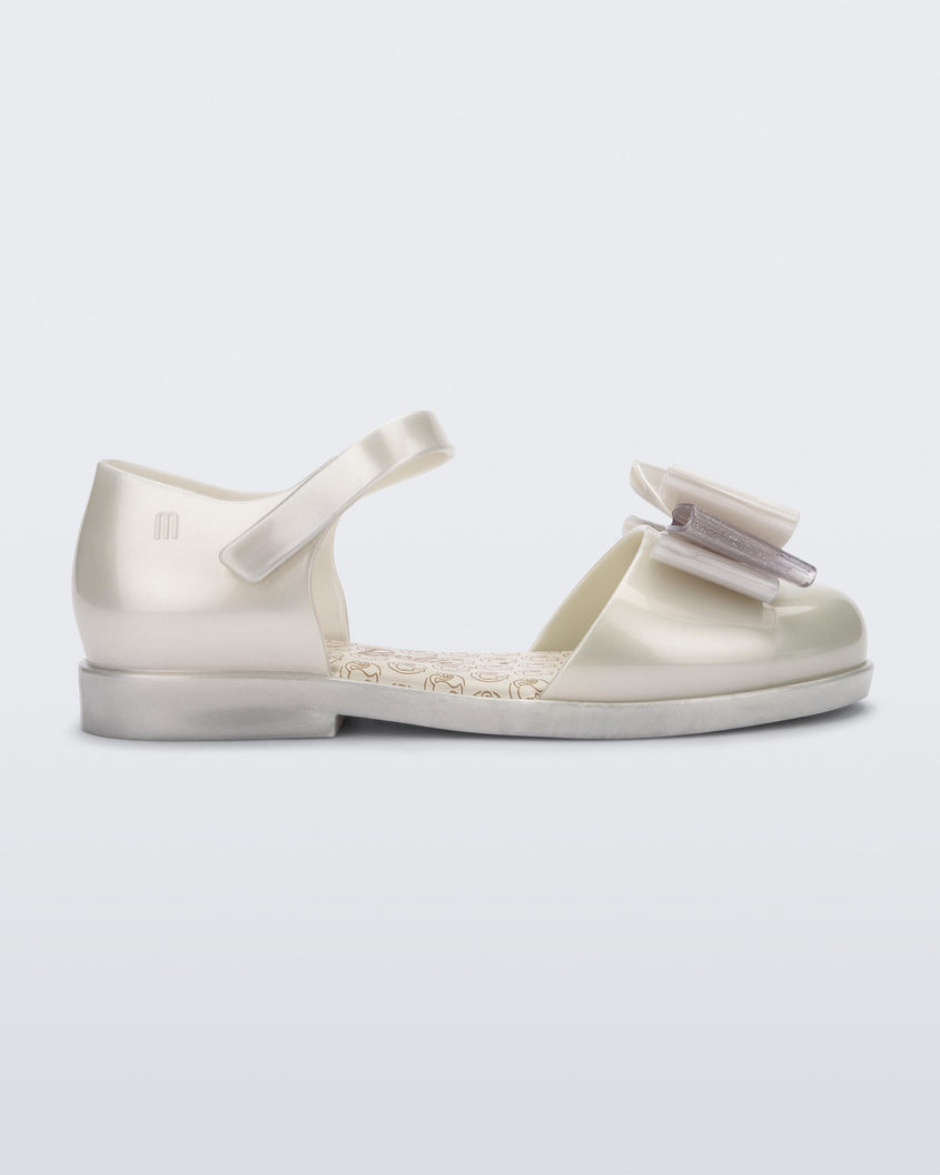 Side view of a metallic white Mini Melissa sandal with a Barbie bow detail on the front toe, metallic white ankle strap and a Barbie logo sole