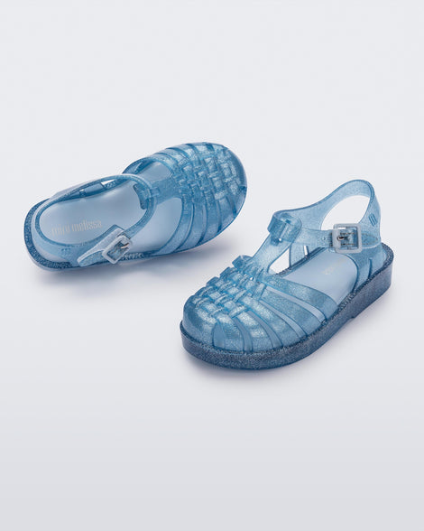 Angled view of a pair of Mini Melissa Possession baby sandals in glitter blue with velcro buckle closure on the ankle straps
