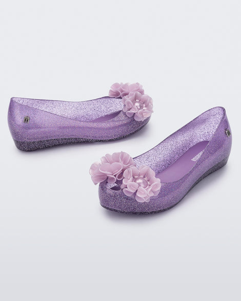 Angled view of a pair clear glitter purple Ultragirl Springtime kids flat with two purple flowers and peep toe.