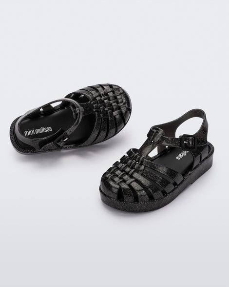 Angled view of a pair of Mini Melissa Possession baby sandals in glitter black with velcro buckle closure on the ankle straps