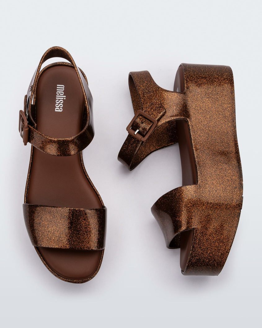 An angled top and side view of a pair of bronze Melissa platform sandals with a front and ankle strap
