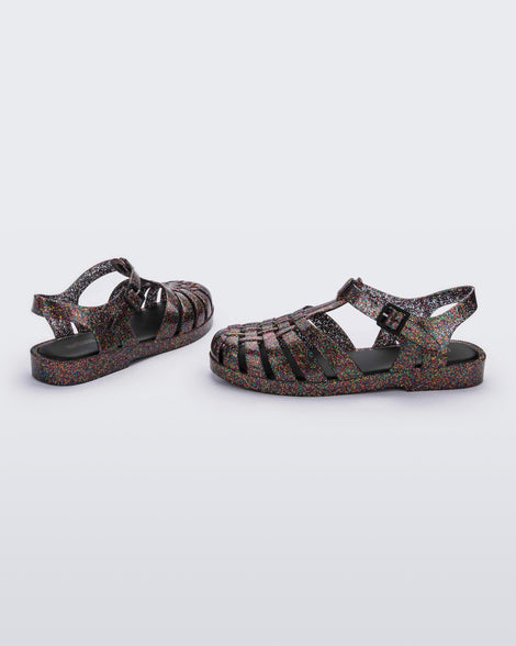 Angled and side view of a pair of multicolored glitter Melissa Possession sandals with a closed toe front weft design connected to a top strap with a buckle.