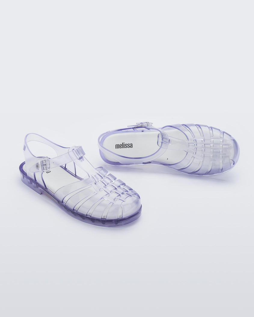 An angled side and top view of a pair of clear Melissa Possession sandals with a closed toe front weft design connected to a top strap with a buckle.