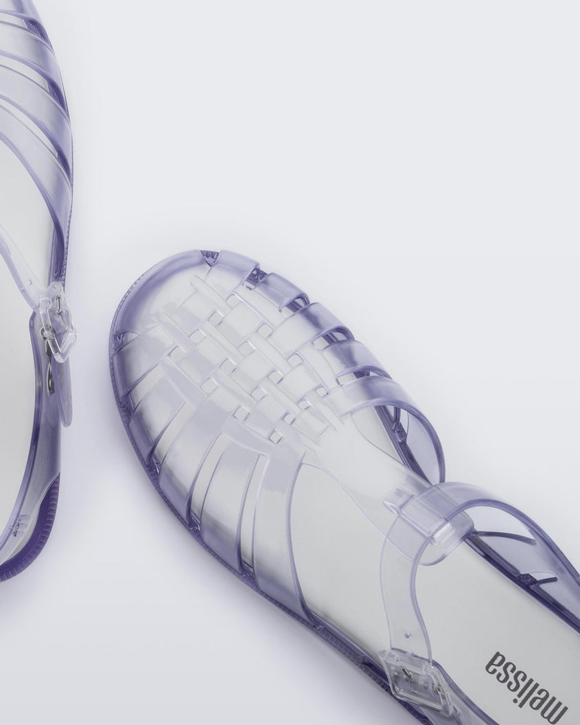 A zoomed in top view of a pair of clear Melissa Possession sandals with a closed toe front weft design connected to a top strap with a buckle.