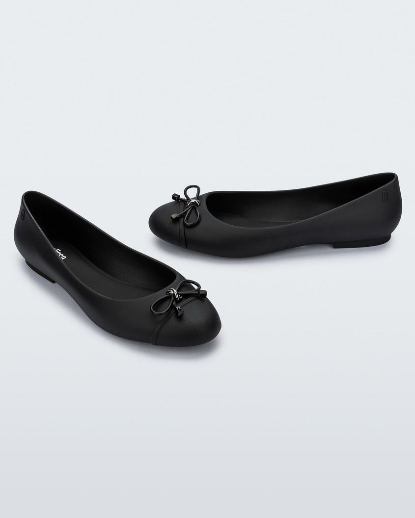 An angled top and side view of a pair of black Melissa flats with a black bow detail with metallic accents on the toe