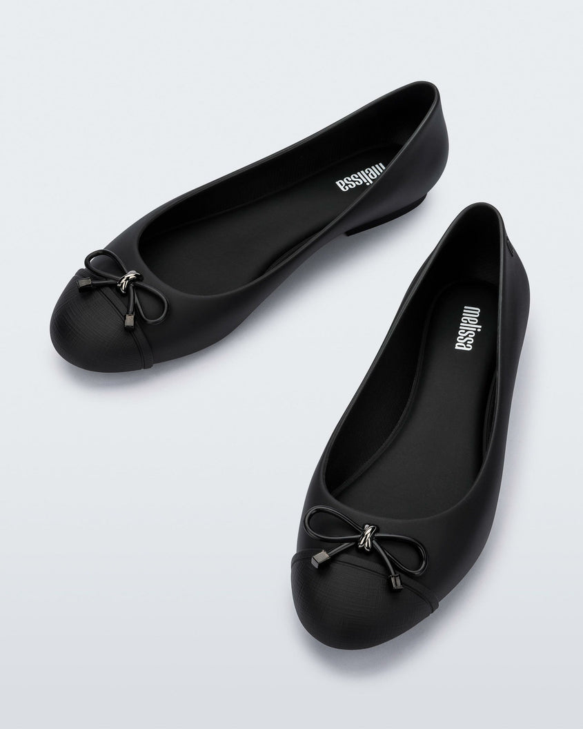 An angled top view of a pair of black Melissa flats with a black bow detail with metallic accents on the toe