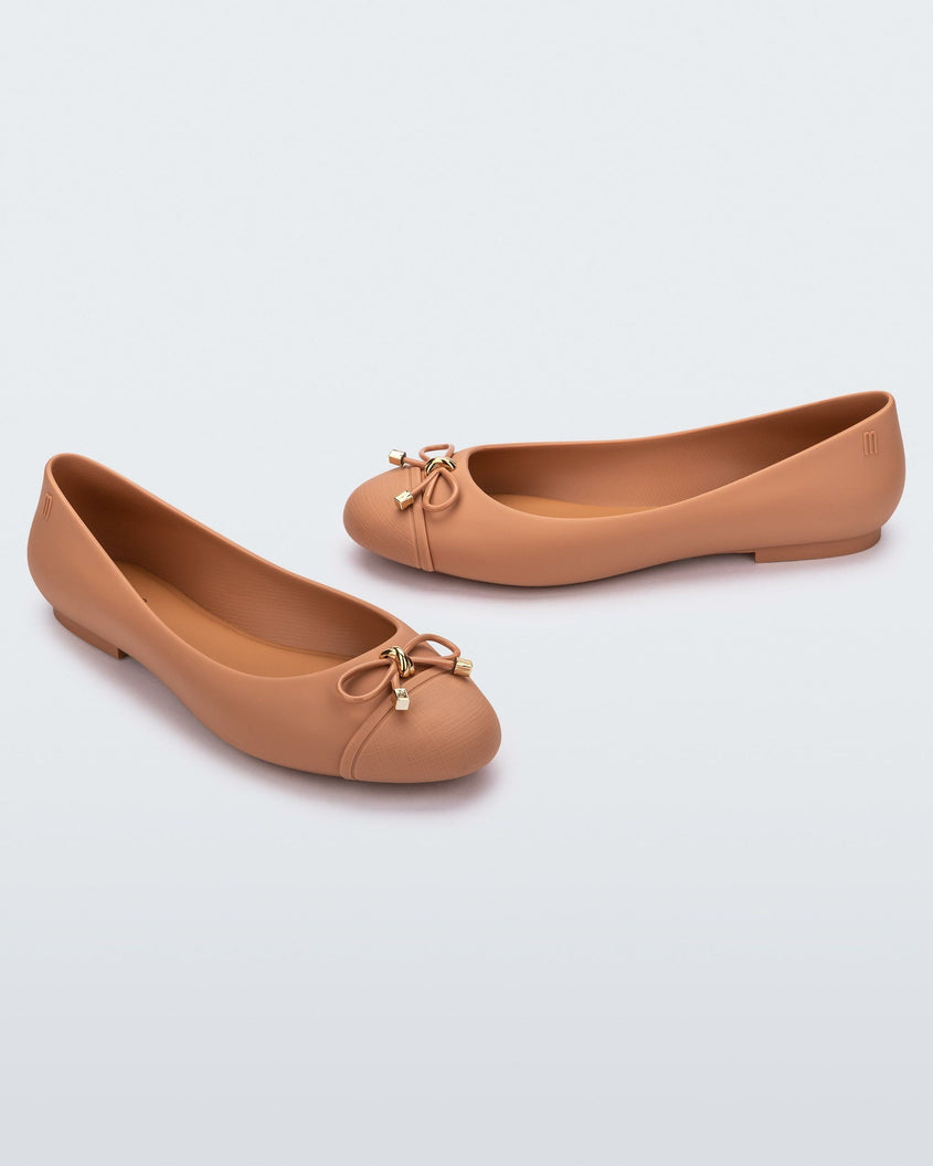 An angled front and side view of a pair of dark beige Melissa flats with a beige bow detail with gold accents on the toe
