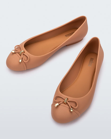An angled top view of a pair of dark beige Melissa flats with a beige bow detail with gold accents on the toe