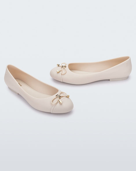 An angled top view of a pair of light beige Melissa flats with a beige bow detail with gold accents on the toe